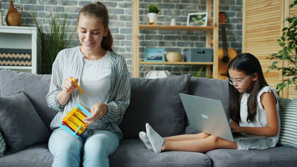 A mother working on a laptop on a sofa while her daughter plays beside her