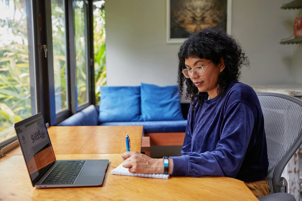 A woman writing notes beside her laptop at a table